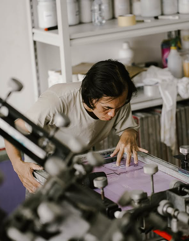 Proses sablon manual kaos menggunakan screen printing dengan tinta ungu di studio percetakan.